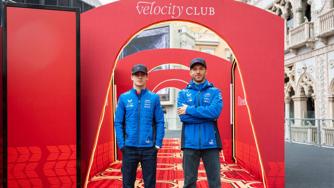 Two people wearing blue jackets standing under a red archway labeled “Velocity Club,” with patterned flooring and European-style architectural details in the background.