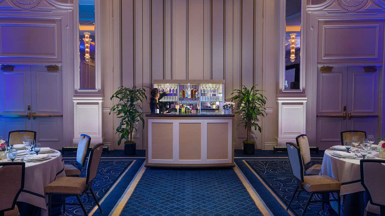 A ballroom setup featuring a central bar with illuminated shelves, two potted plants, round banquet tables with chairs, and elegant wall detailing under soft blue lighting.