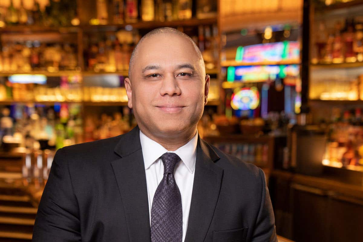 A man with a shaved head is smiling slightly. He is wearing a dark suit, white shirt, and a dark patterned tie, and is leaning against a bar with a warmly lit backbar.