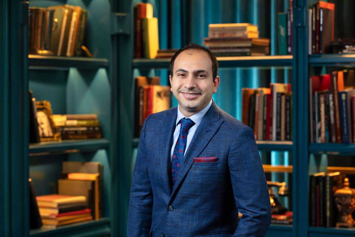 A professional headshot of a smiling man with short dark hair, wearing a textured blue suit with a patterned blue tie and a red pocket square. He is standing in front of a blue bookshelf filled with books and vintage decor.