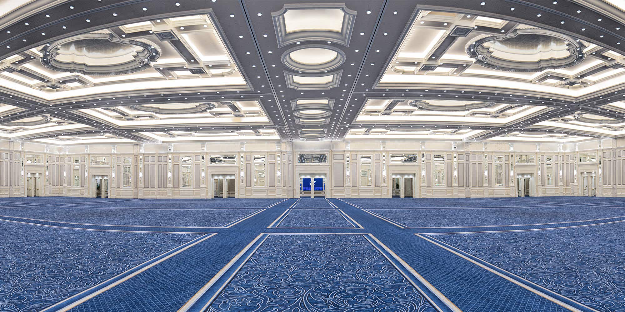 Spacious empty ballroom with ornate white detailing, blue patterned carpet, high ceilings, recessed lighting, and symmetrical doorways.