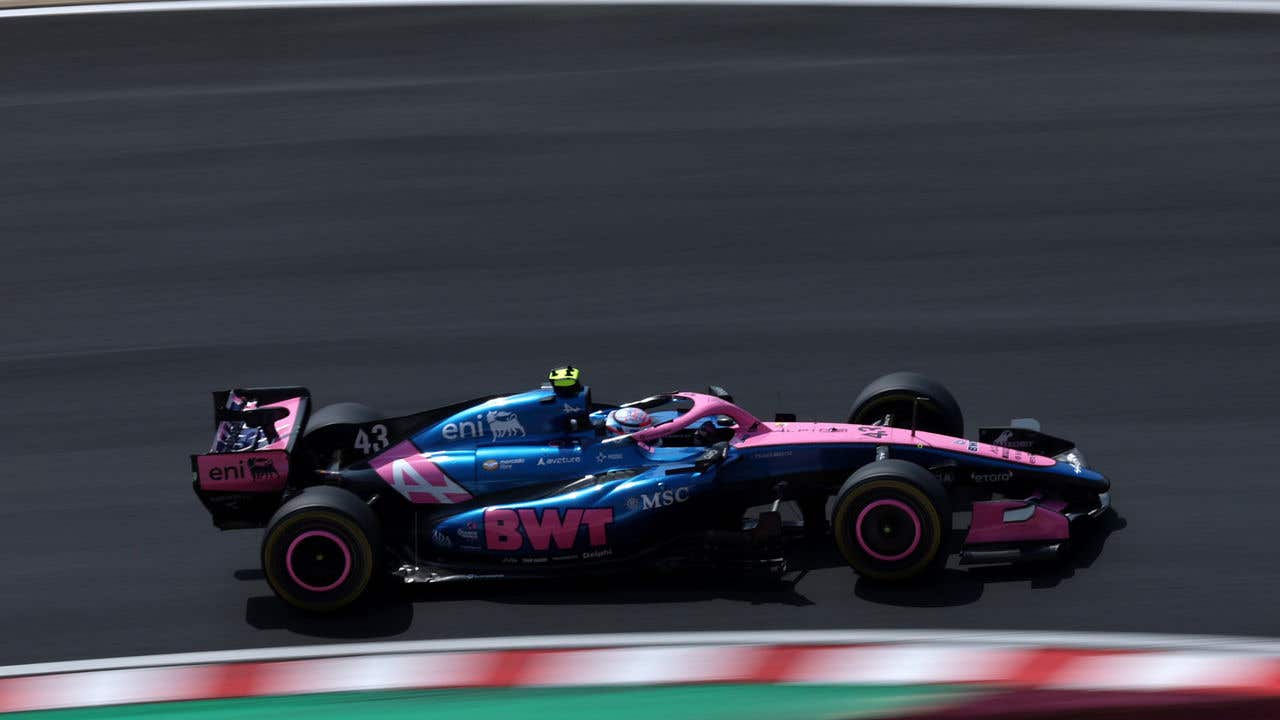 Side view of a pink and blue Formula 1 race car driving at speed on a racetrack, with motion blur on the asphalt and curbing visible along the edge of the track.
