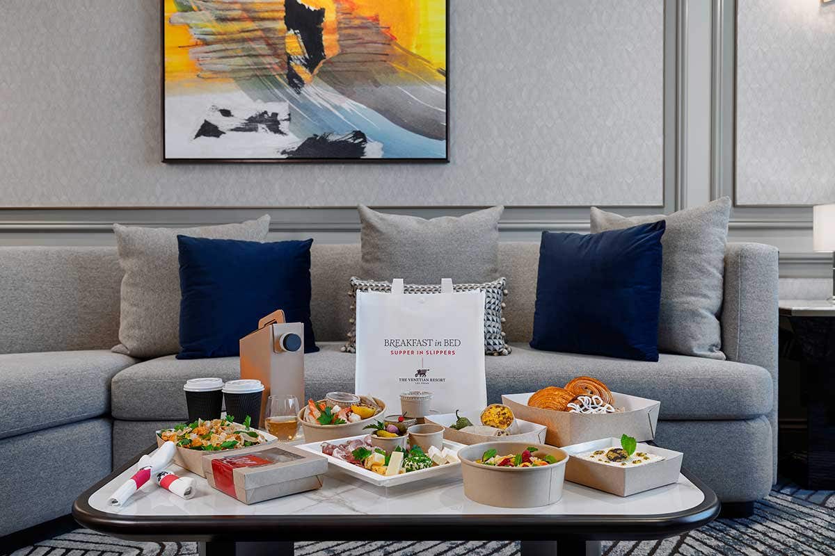 A spread of food in to-go containers sits on a coffee table in a hotel living room in front of a couch.