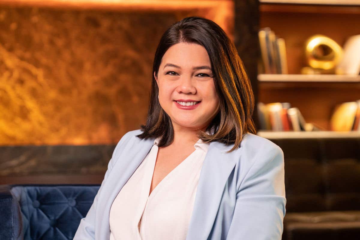 A woman with shoulder-length dark hair, smiling, wearing a white blouse and a light blue jacket, is seated on a blue chair in a warmly lit room.