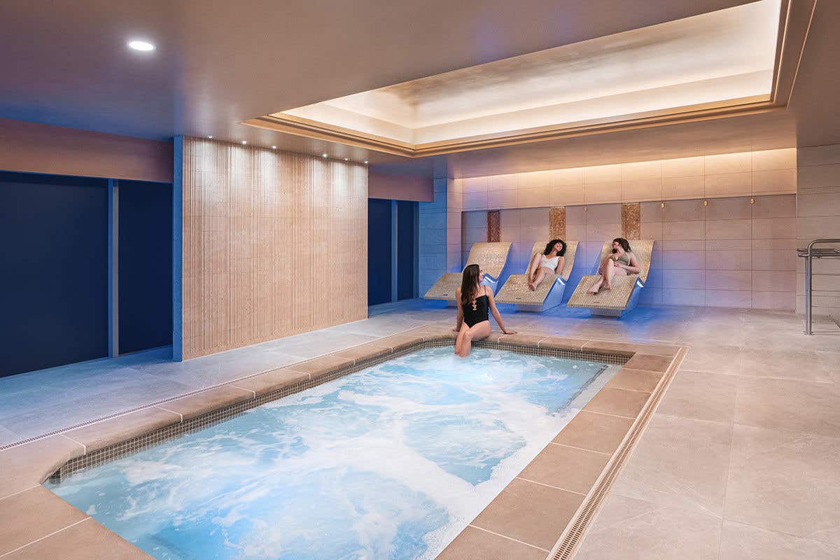 Three women relax in and around a bubbling indoor whirlpool. One woman in a black swimsuit is sitting on the edge, dipping her feet in the water, while two other women lounge on cushioned chairs in the background. The spa area has a serene design with tiled walls, soft backlighting, and a modern, recessed ceiling light, creating a calm and inviting environment.
