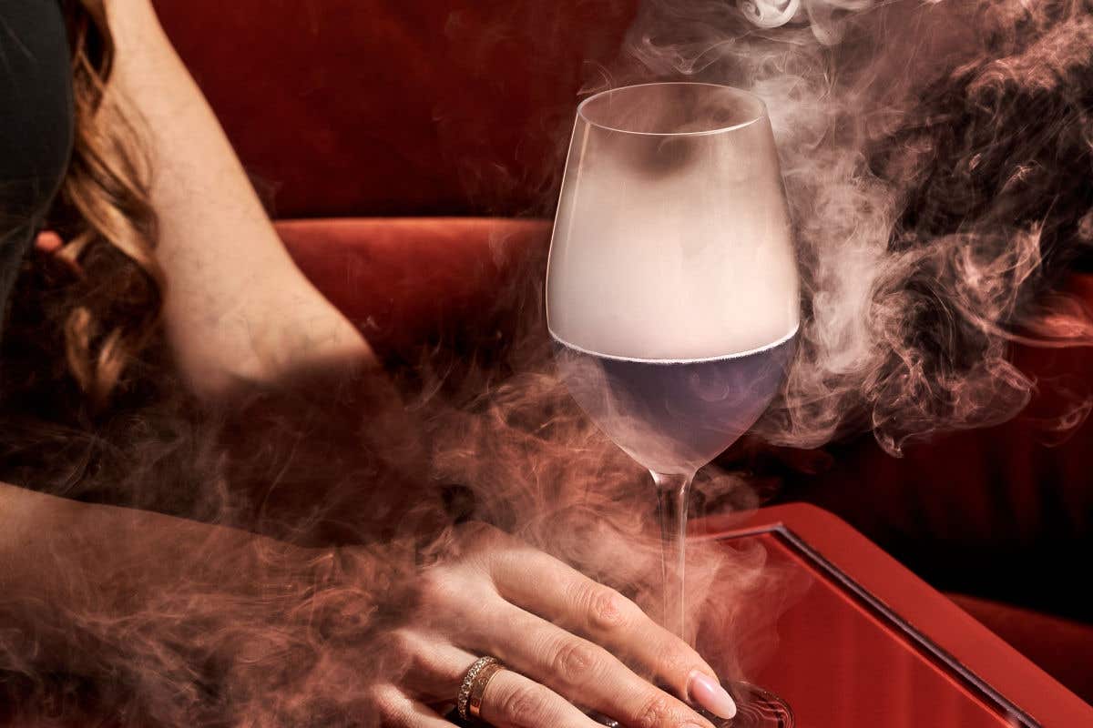 A close-up of a person's hand holding a wine glass filled with a dark liquid and billowing white smoke, set against a plush red backdrop.
