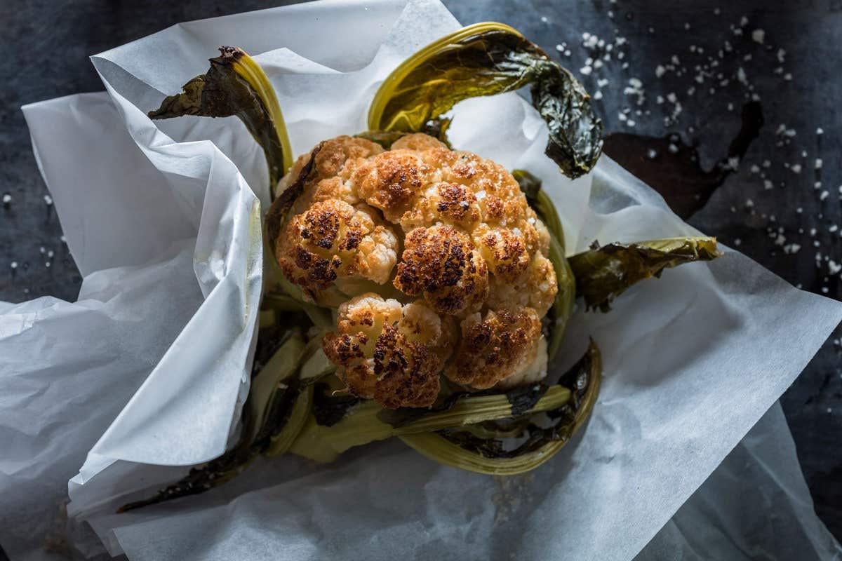 Roasted cauliflower on parchment paper with charred leaves on a dark background, sprinkled with sea salt.