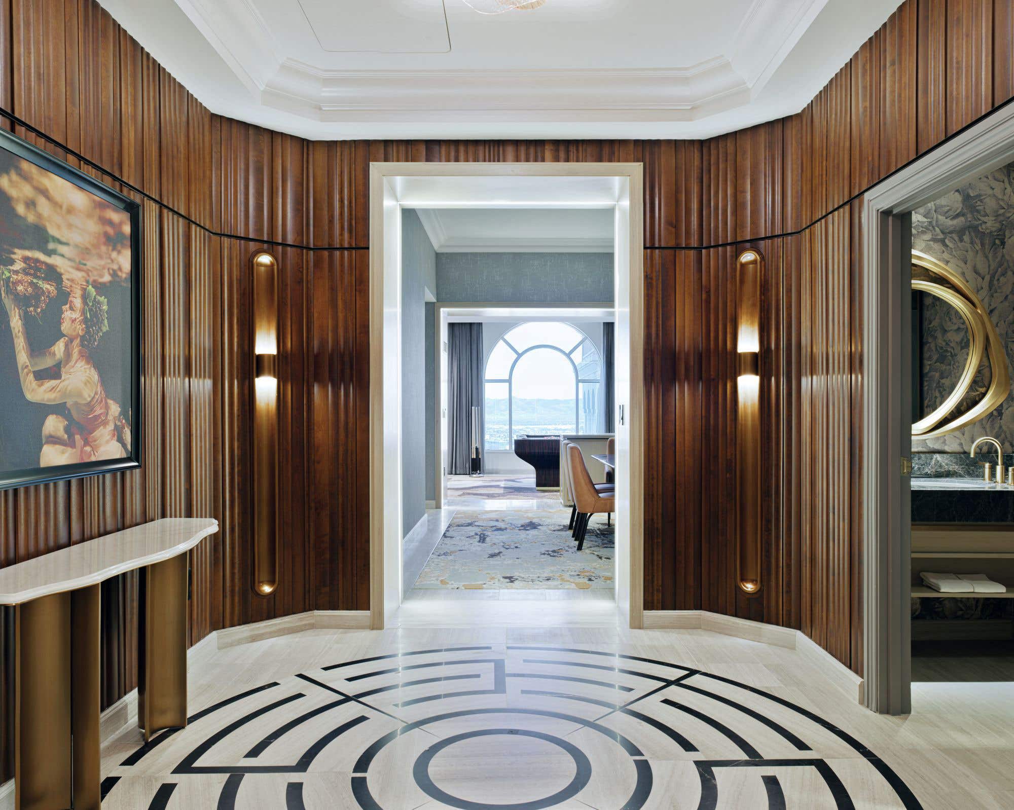 Luxurious suite hallway with curved wood‑paneled walls, art deco lighting, patterned marble floor, and a view into a sitting area and bathroom beyond.